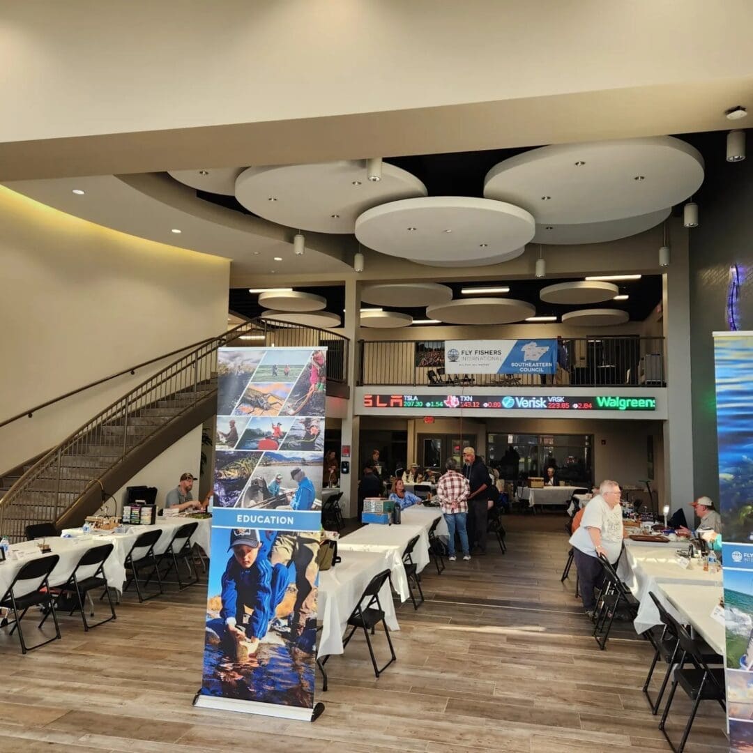 A spacious indoor event area featuring tables with white tablecloths and chairs. Attendees are gathered near promotional banners and booths. A stairway and upper balcony are visible in the background.