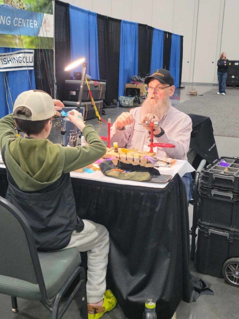 A bearded man and a young boy sit at a table with fishing equipment, apparently engaged in fly tying. They are in a convention or exhibition hall with a banner and cases visible in the background.