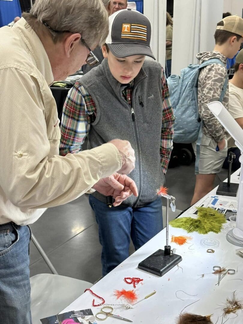 An older man instructs a boy in a cap on tying a fishing fly at a table covered with fly-tying materials and tools.