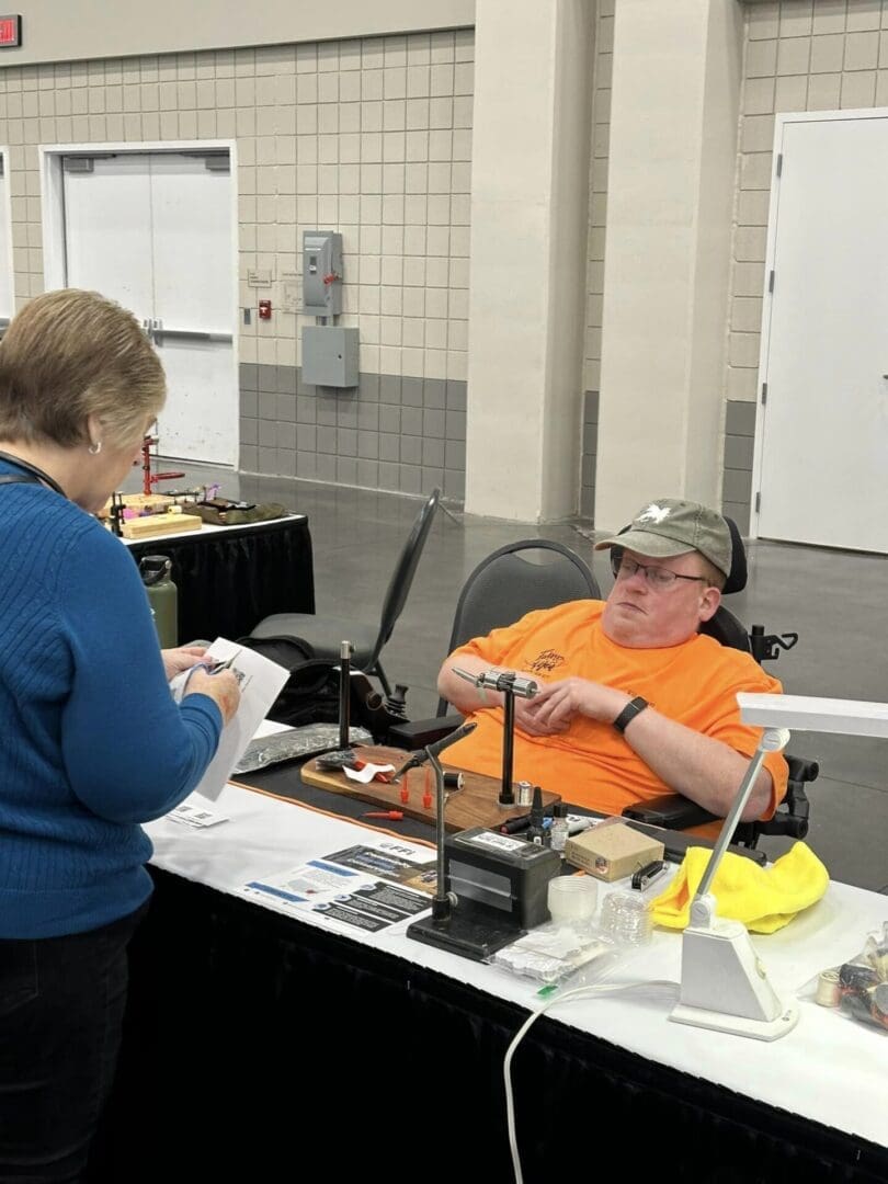 A man sitting at a booth demonstrates a craft to a woman who is taking notes at an indoor event. Various tools and materials are arranged on the table.