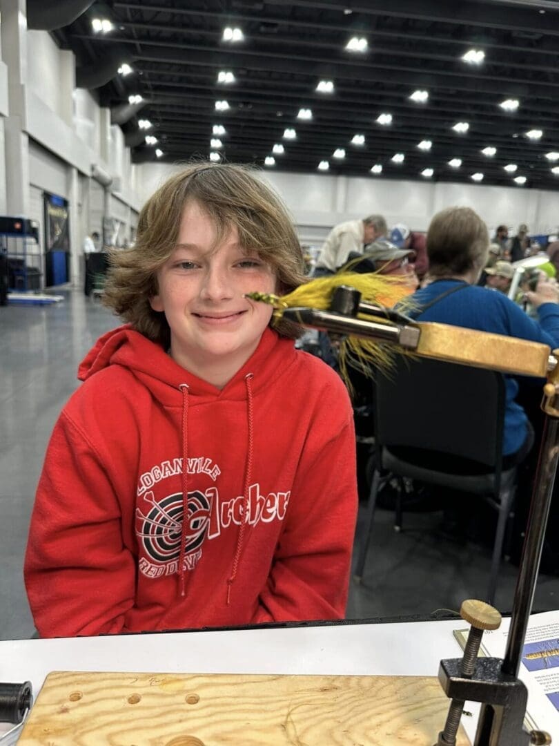 A young person in a red hoodie sits at a table inside a large hall, next to fly-tying equipment with a fly attached to a vise. People are seated and standing in the background.