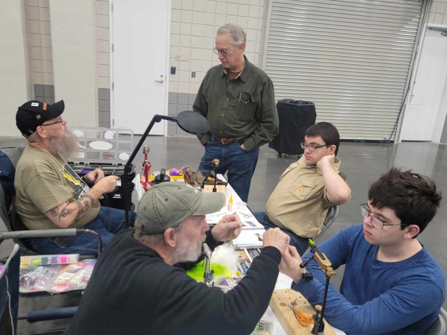 A group of five people are seated around a table, engaged in an activity. Some individuals are tying flies, while others observe. One person stands nearby, watching.