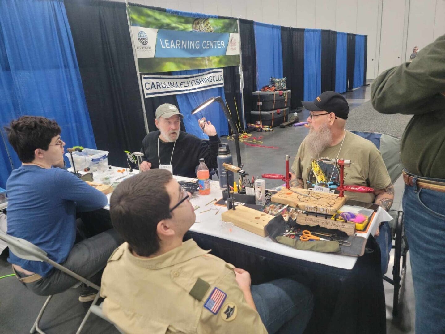 Four individuals are seated at a table with fly fishing equipment, engaging in conversation. Behind them is a banner that reads 