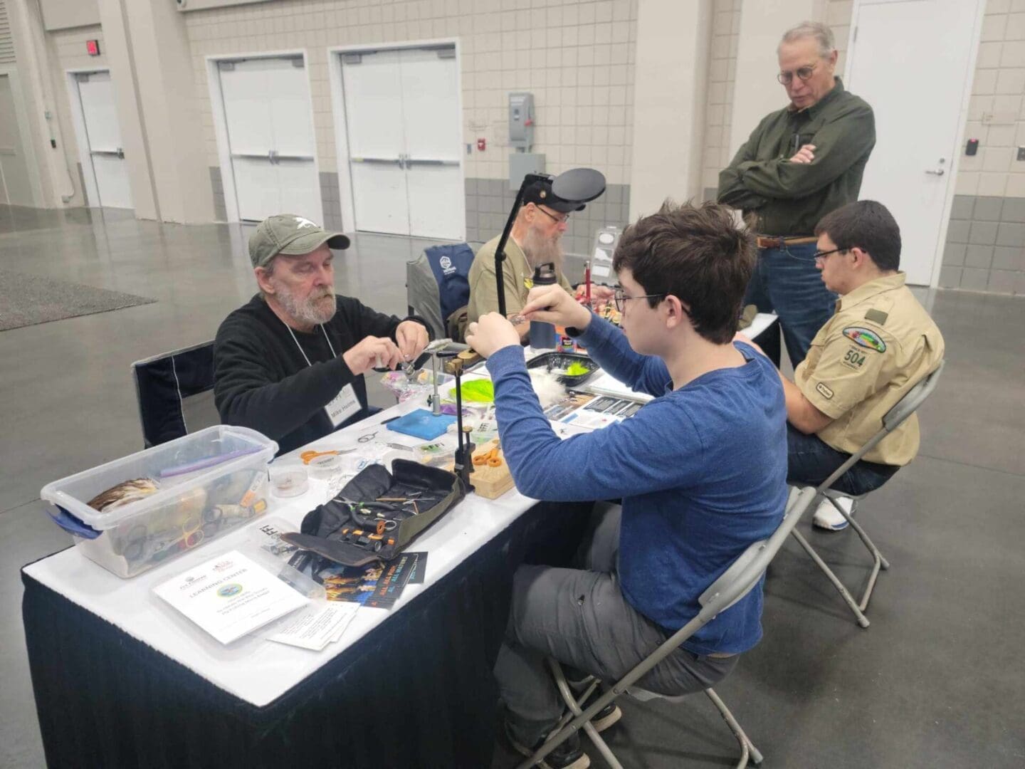 Four individuals are seated at a table, engaging in an activity involving various tools and materials. Two of them are in scout uniforms. An older man stands nearby observing.