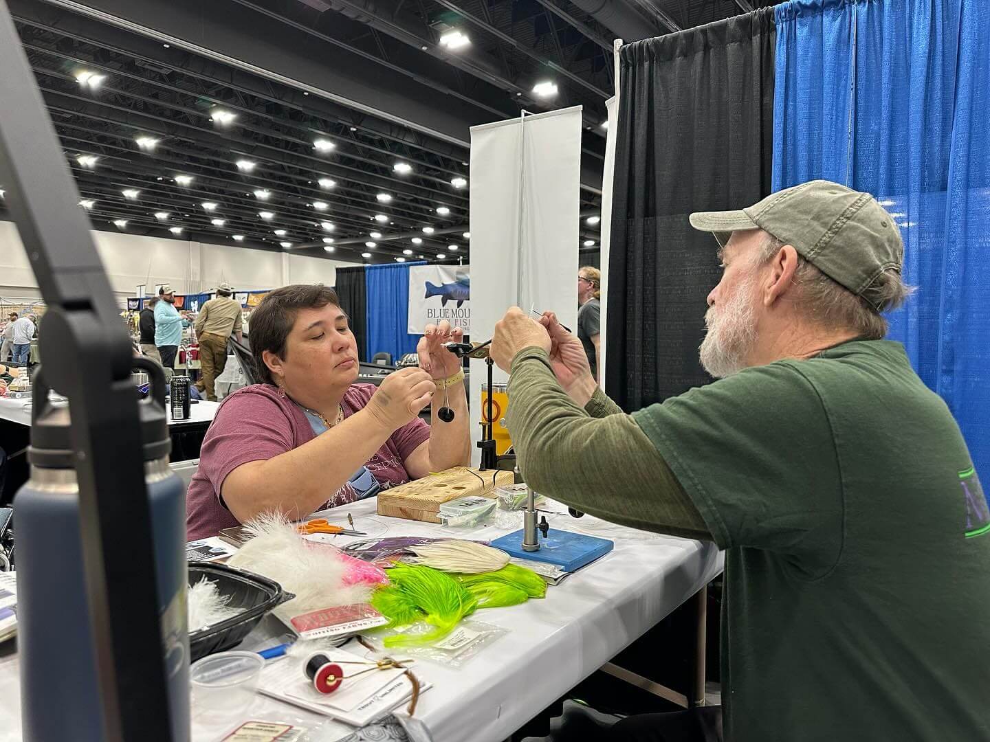 Two people at a table in a convention center are tying flies. The table is covered with tools and materials such as feathers and threads used for fly tying.