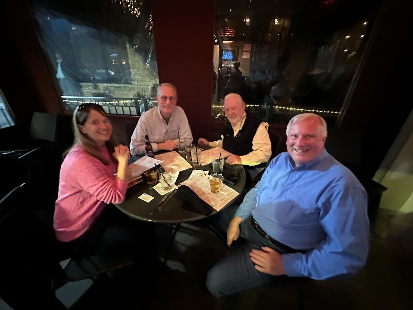 Four people are seated around a table in a dimly lit restaurant, smiling at the camera. Glasses and menus are on the table.