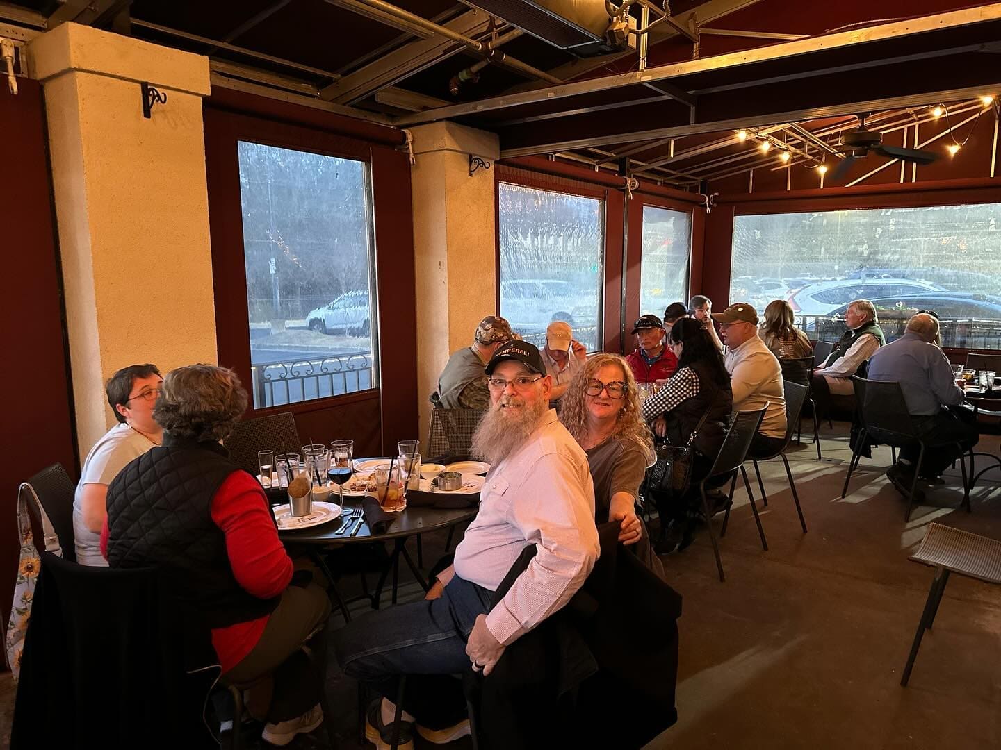A group of people sits at a long dining table in a restaurant with large windows. Two people in the foreground, wearing glasses and smiling, pose for the camera.