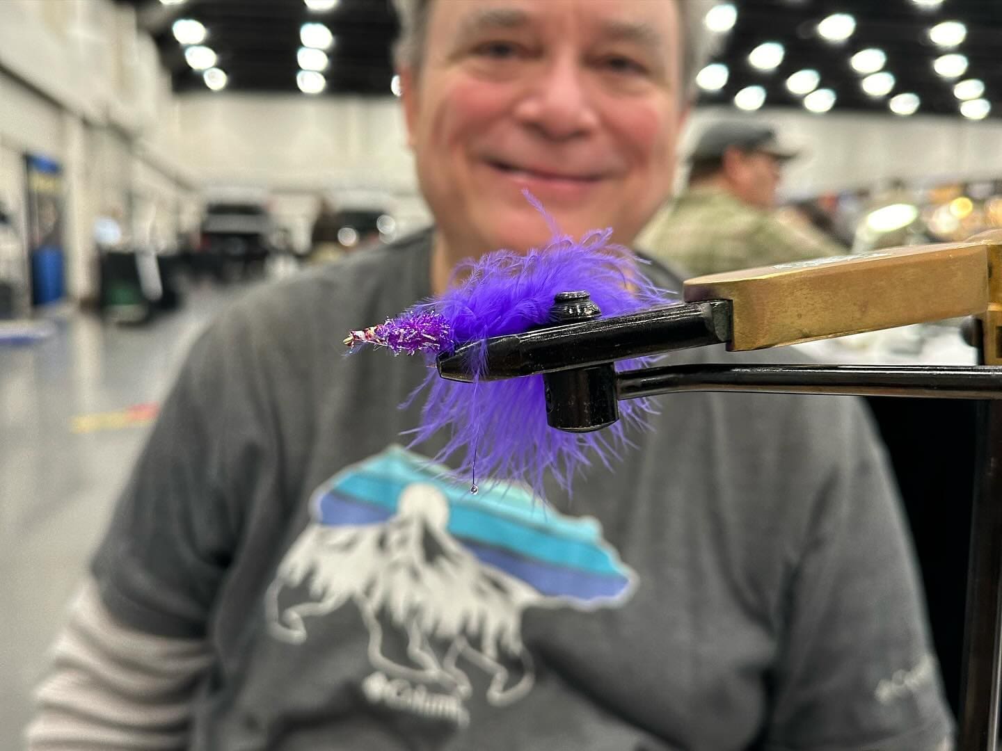 A person is sitting and smiling in the background while a close-up focus is on a purple feathered fly fishing lure held in a vise in the foreground.