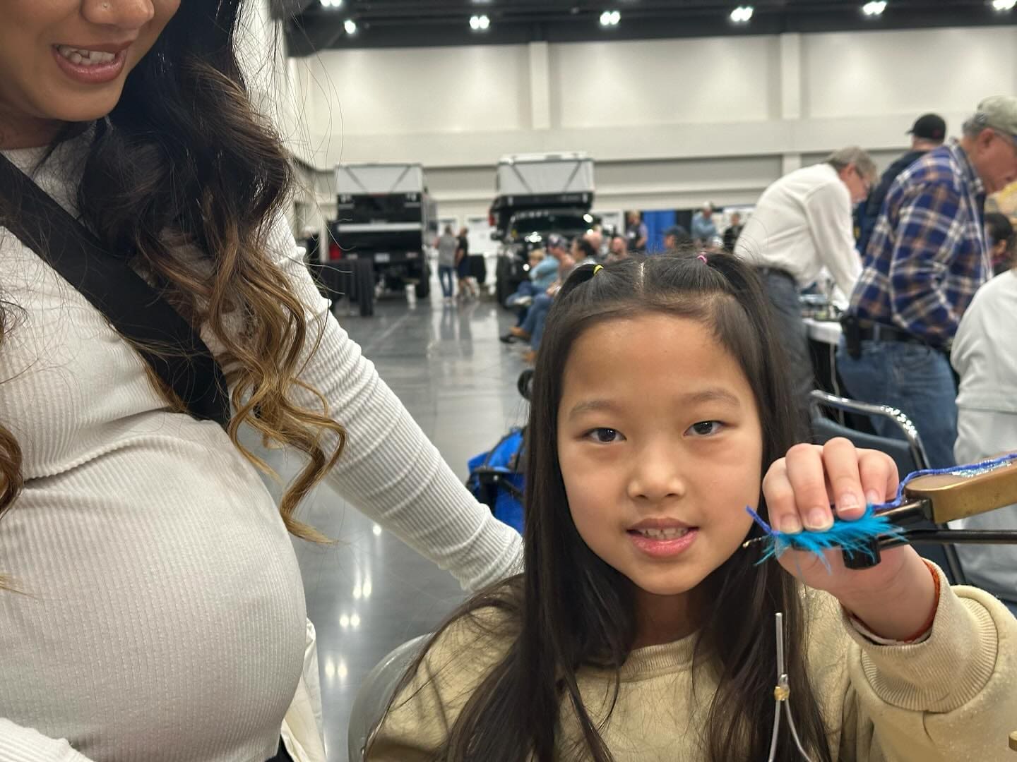 A young girl holds up a blue feather while sitting at a table in a large indoor space. An adult woman stands beside her, and other people are visible in the background.