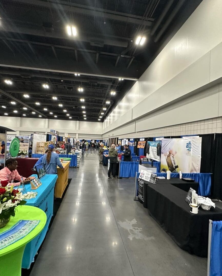 Wide view of a convention hall with various booths set up on both sides. People are seen walking and browsing the displays at the event.