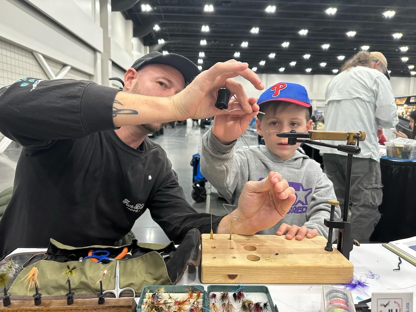 An adult and child are tying fishing lures at a table filled with fly-tying tools and materials. They are intently focused, working together in an indoor setting.