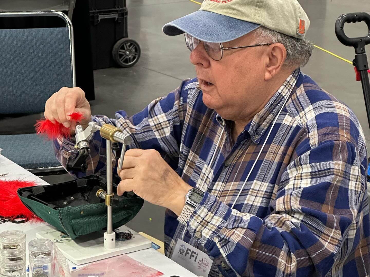 A man in a plaid shirt and cap is sitting at a table, assembling a lure with red feathers, using tools and materials. He wears a lanyard with 