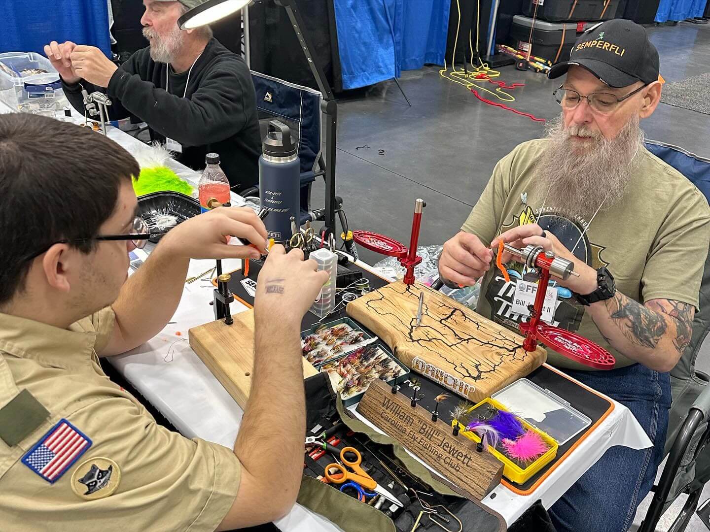 Two individuals sit at a table filled with tools and supplies, engaged in fly tying. One person wears a scout uniform while the other has a long beard and glasses. They are inside a convention hall.
