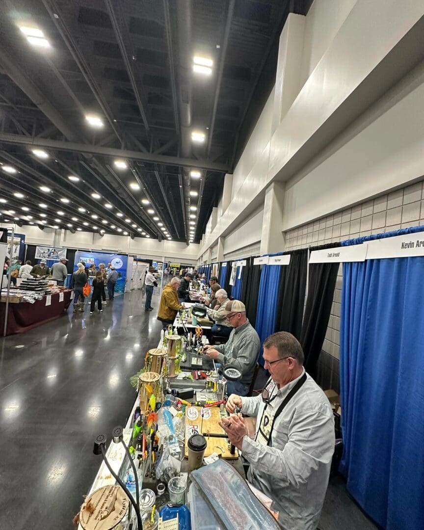A group of people are seated at tables working on projects at an indoor event. Banners and booths are visible in the background.