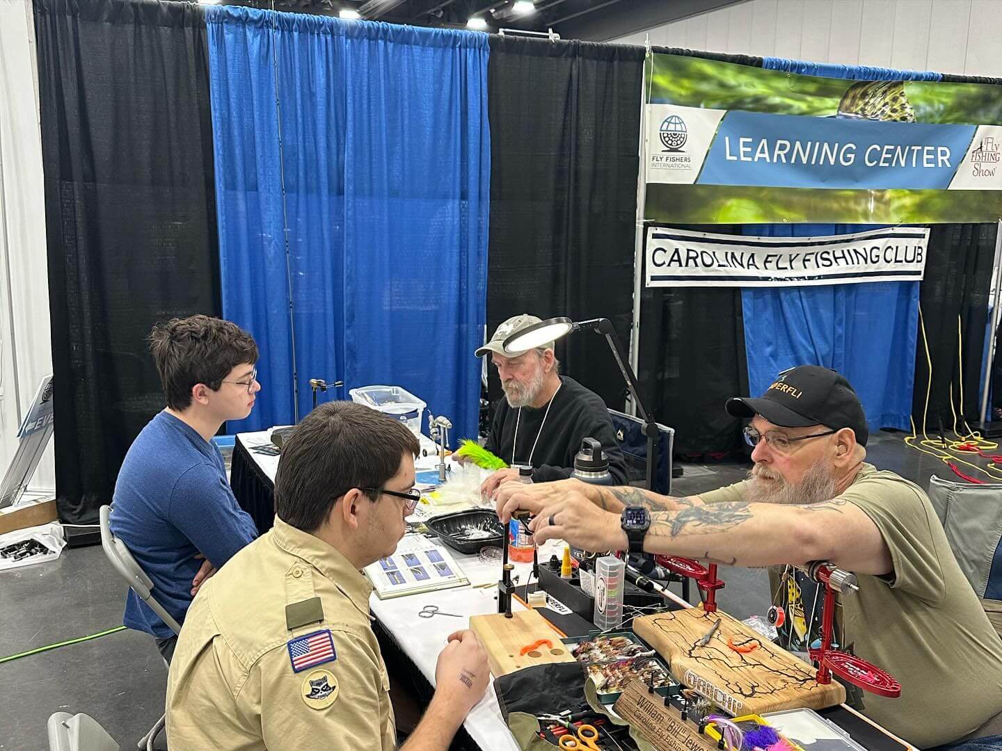 Two men in scouting uniforms and two older men work on fly-tying projects at a table in a booth labeled 