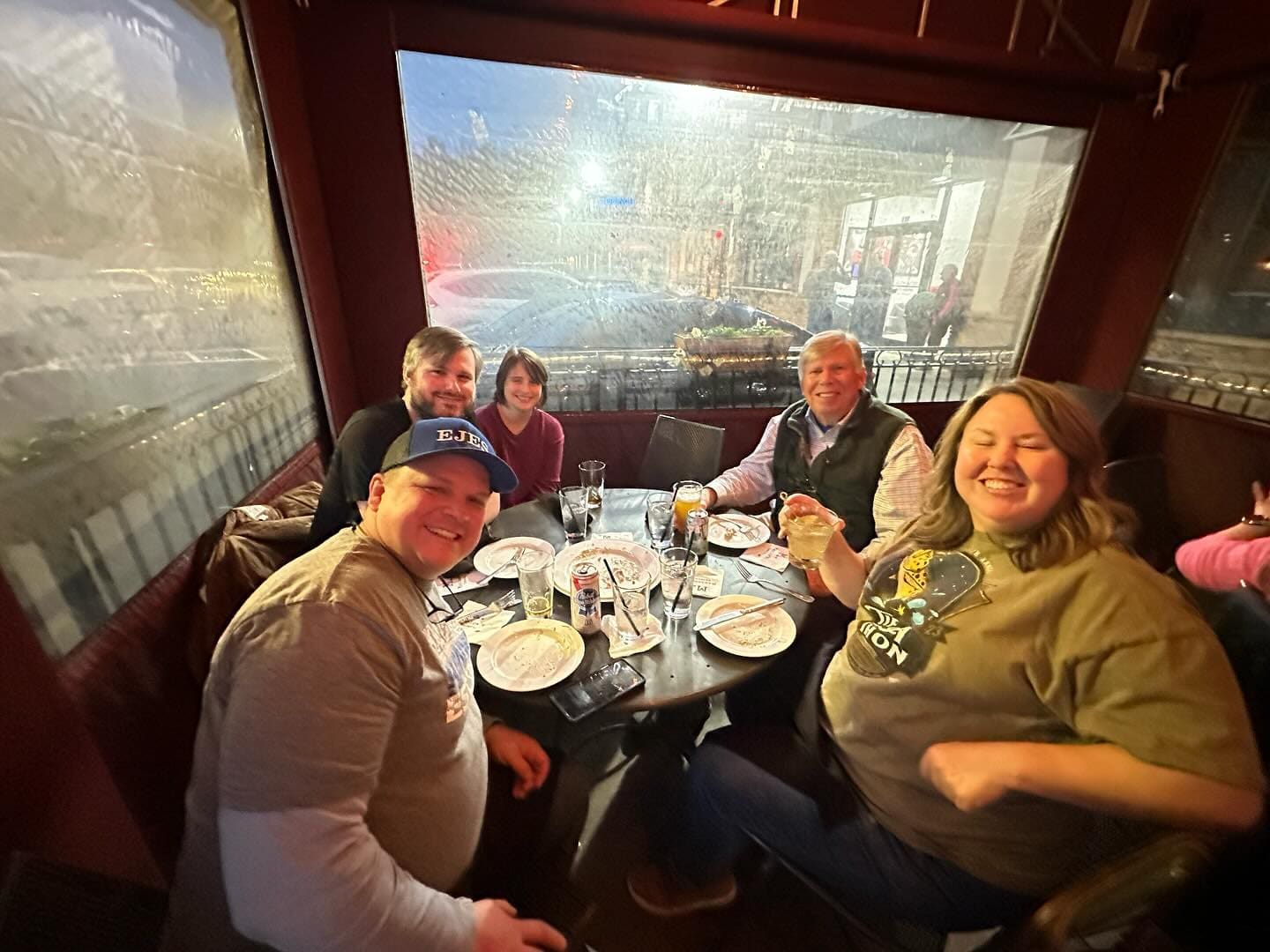A group of five adults smiling and posing for a photo while seated around a table at a restaurant, with food and drinks on the table in front of them. The restaurant has large windows in the background.