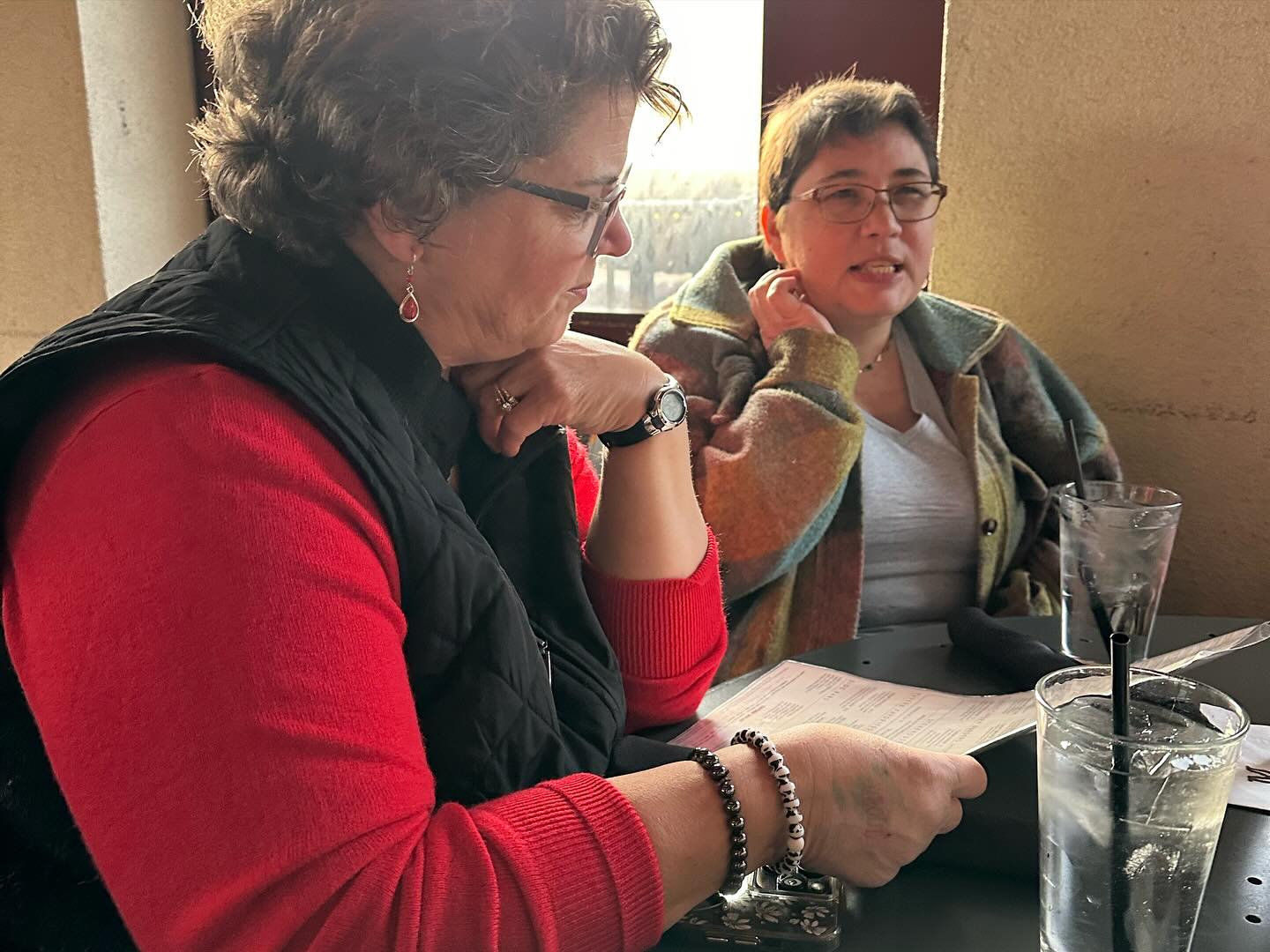 Two women sitting at a table in a restaurant. One is reading a menu while the other looks on. Glasses of water with straws are on the table.