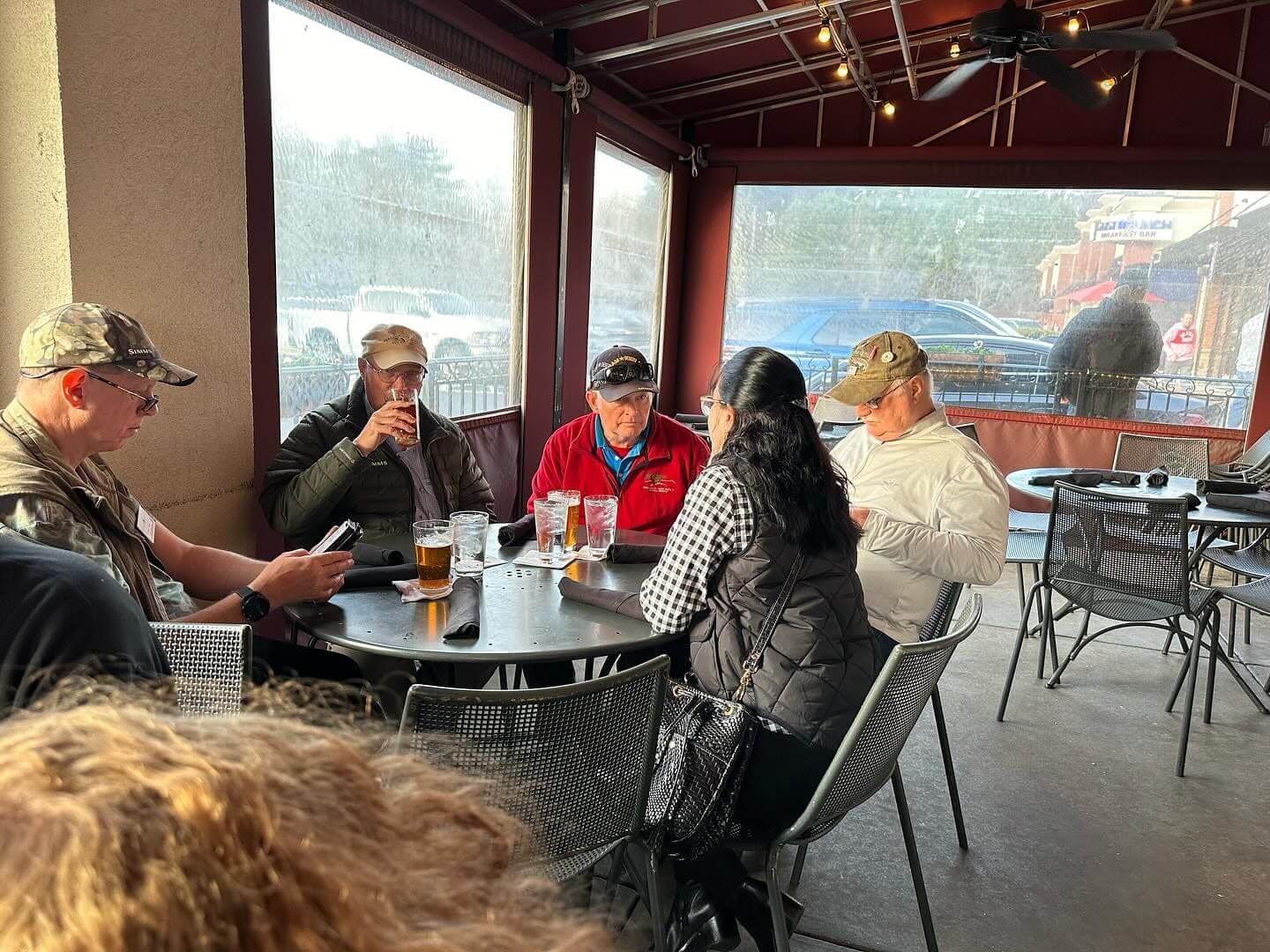 Five people sit around a table on an outdoor patio. Two individuals are drinking beer, and three are engaged in conversation. Jackets and hats appear to be part of their attire.