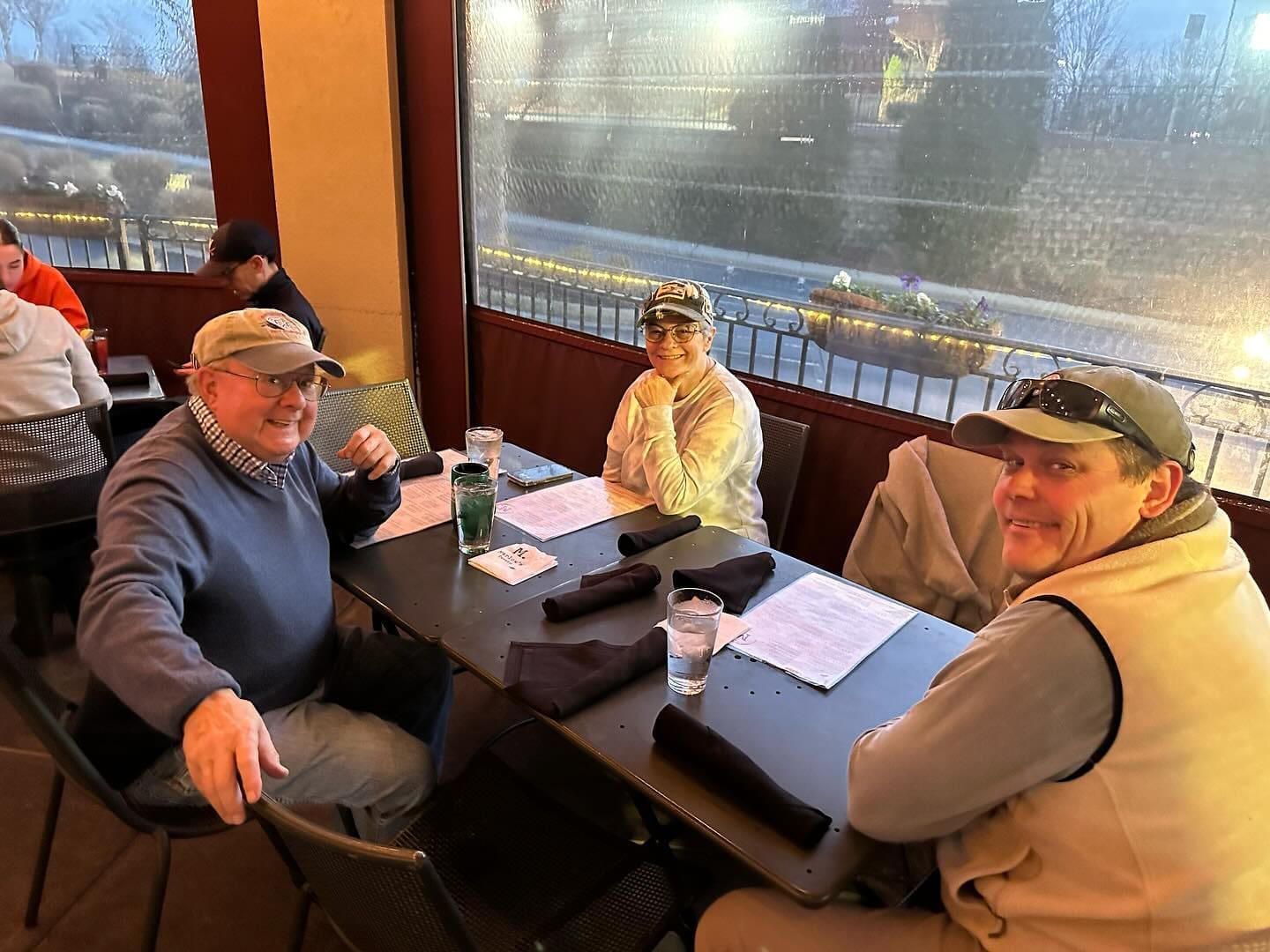 Three people sit at an outdoor café table with menus and drinks, engaging in conversation and smiling at the camera.