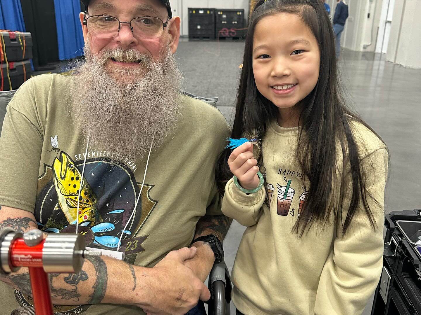 An older man with a long beard and glasses, wearing a graphic T-shirt, smiles beside a young girl holding a small blue feather inside a large indoor venue.