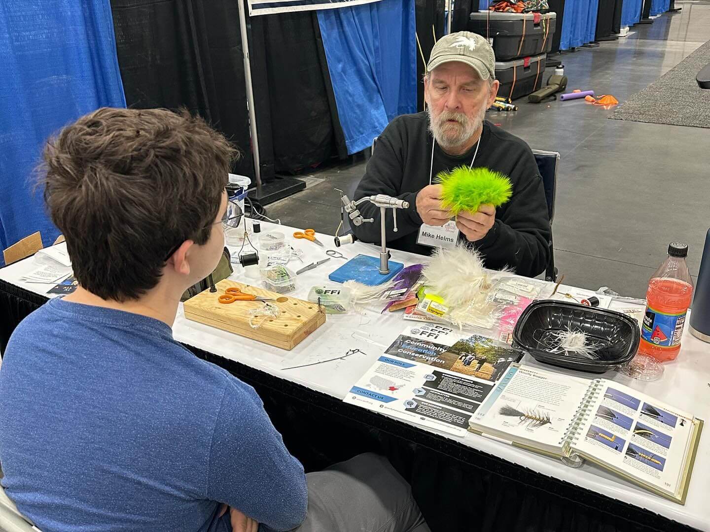 A man is demonstrating fly tying at a booth to a seated man. The table is filled with fly tying materials and tools.