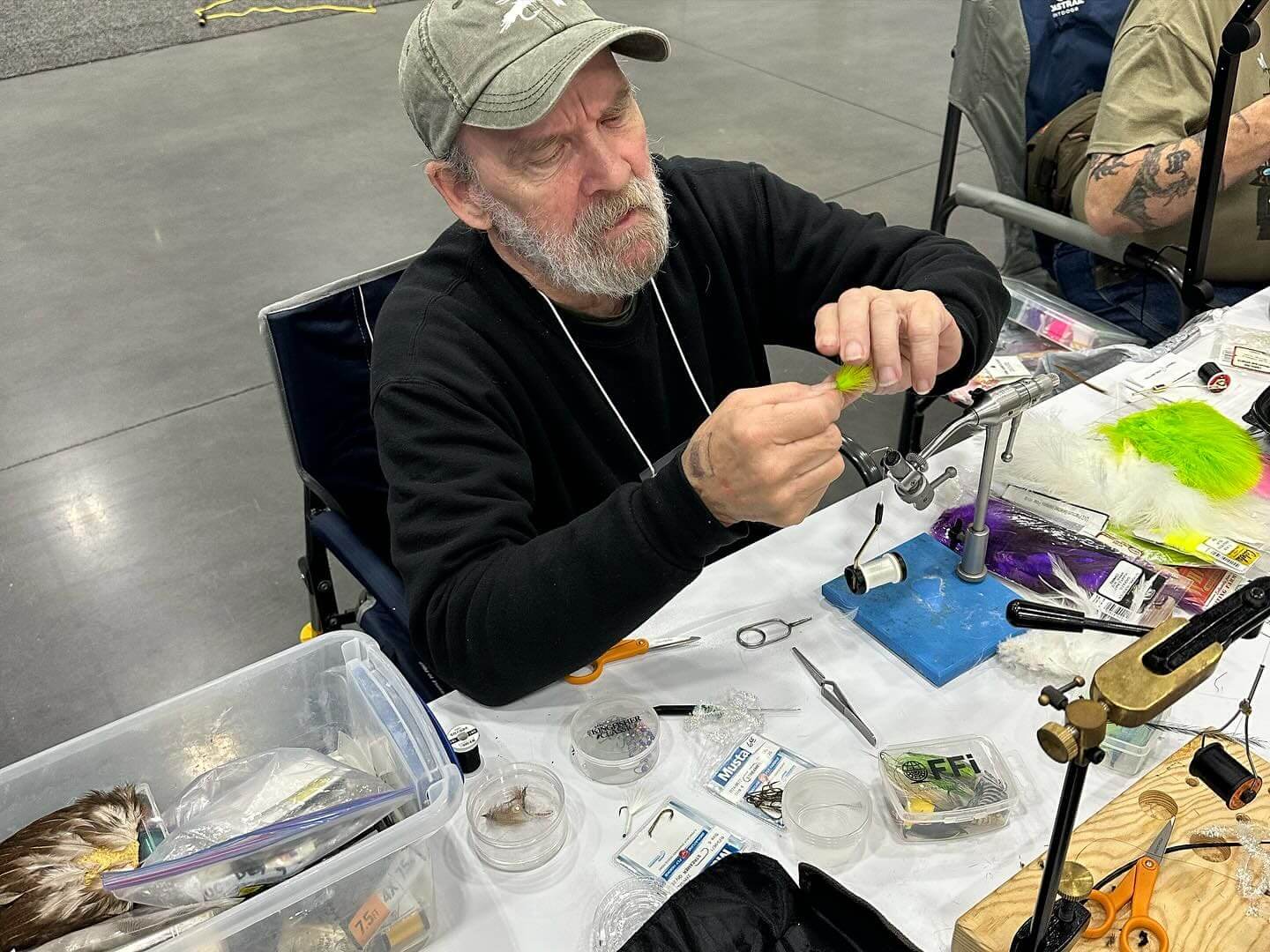 An elderly man with a gray beard sits at a table organizing fly fishing materials and tools. He is holding a fishing lure and appears focused on his task. Various supplies are spread on the table.