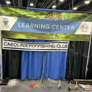 A booth at a convention features banners for the Learning Center, Fly Fishing Show, and the Carolina Fly Fishing Club, with various fishing poles displayed in front.