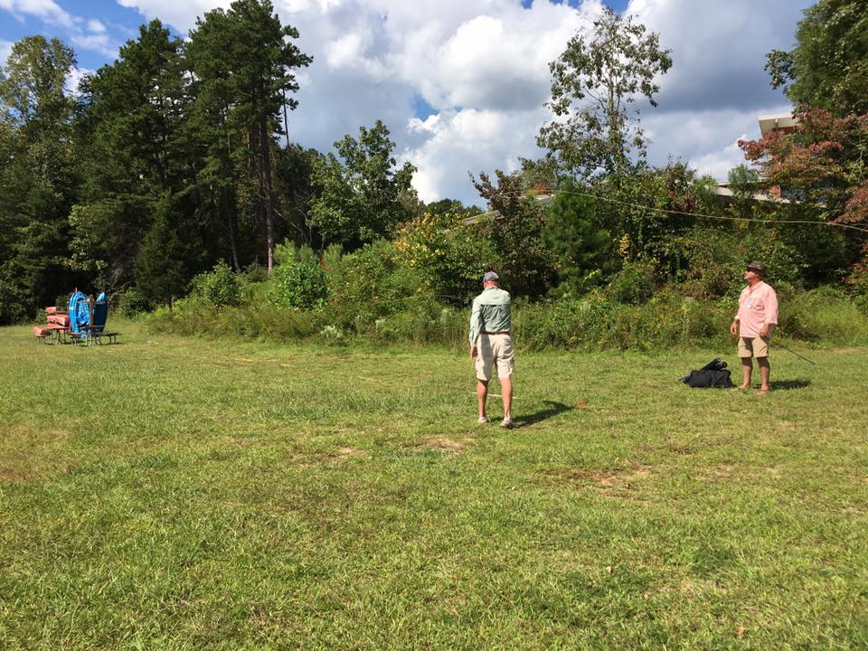 Two men stand on a grassy field with trees in the background. One man faces away, while the other faces towards the camera. Blue chairs and a table are seen in the distance on the left.