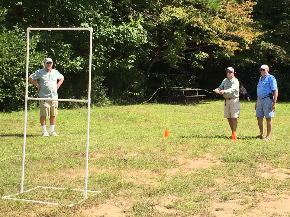 Three men participate in an outdoor casting practice session, using a framework with two orange cones as guides on a grassy field with trees in the background.