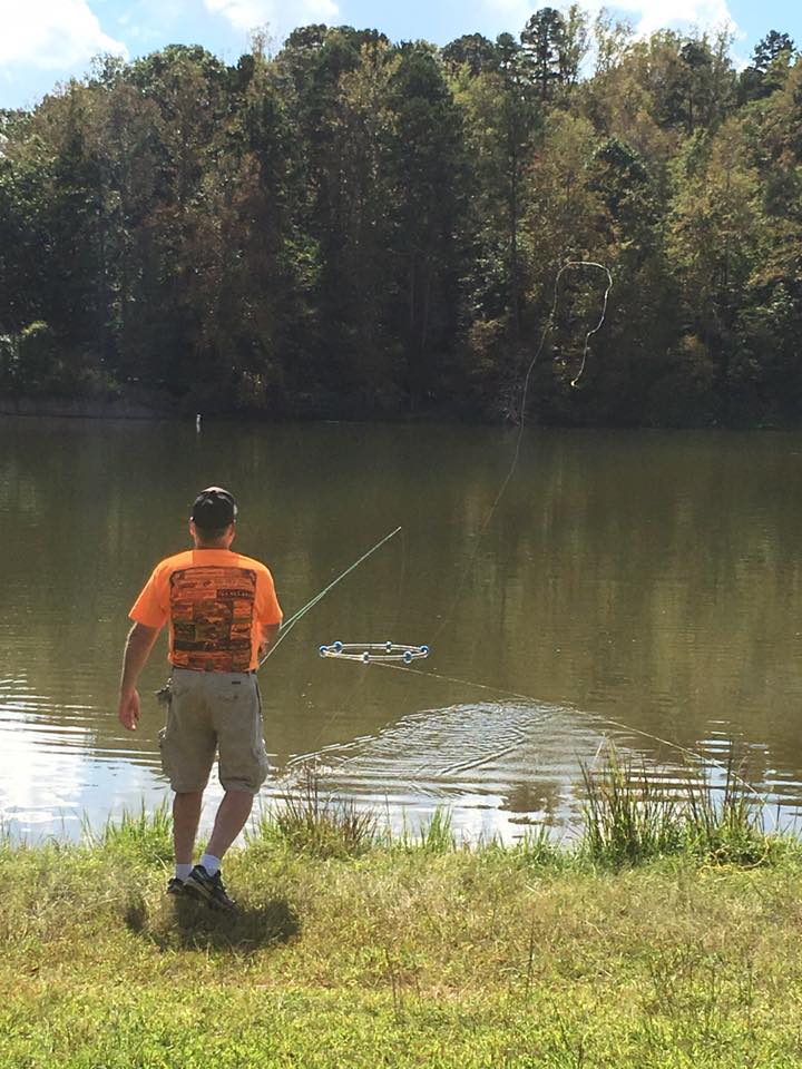 A man wearing an orange shirt and khaki shorts is fishing by a calm lake surrounded by trees, casting a fishing line into the water.