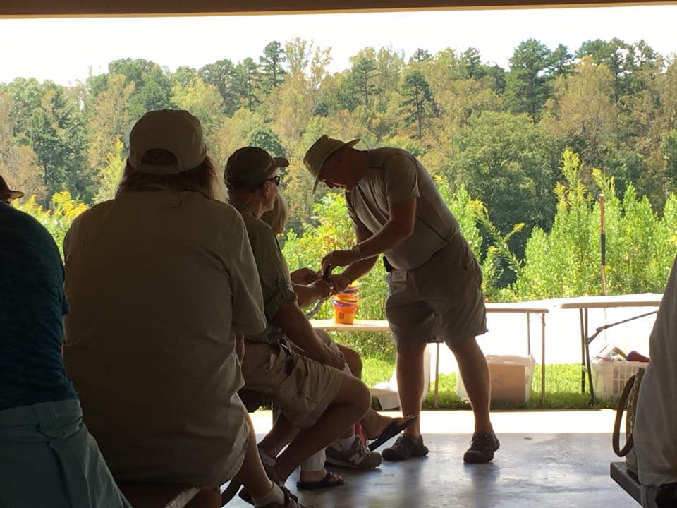 A person wearing a hat demonstrates something to a group seated under a pavilion. Trees and plants are visible in the background.