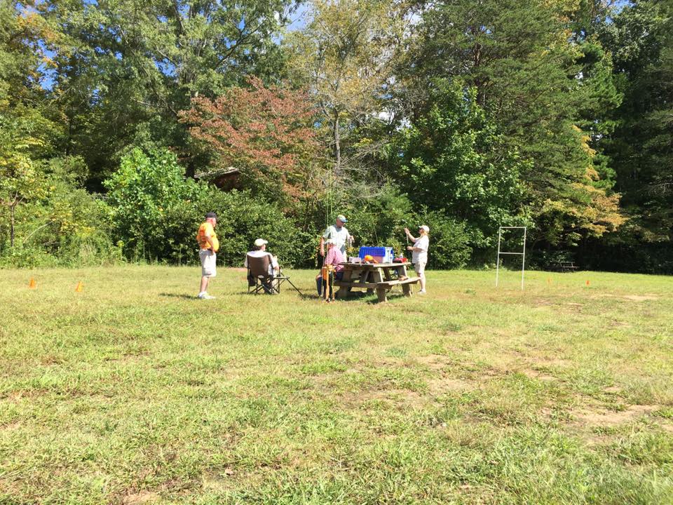 A group of people is gathered around a picnic table in an open grassy area surrounded by trees. Some are seated while others stand, engaging in conversation or activities.