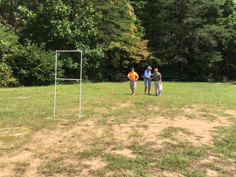 Three people walk across a grassy area with trees in the background. A white frame structure and a yellow hula hoop are on the ground nearby.