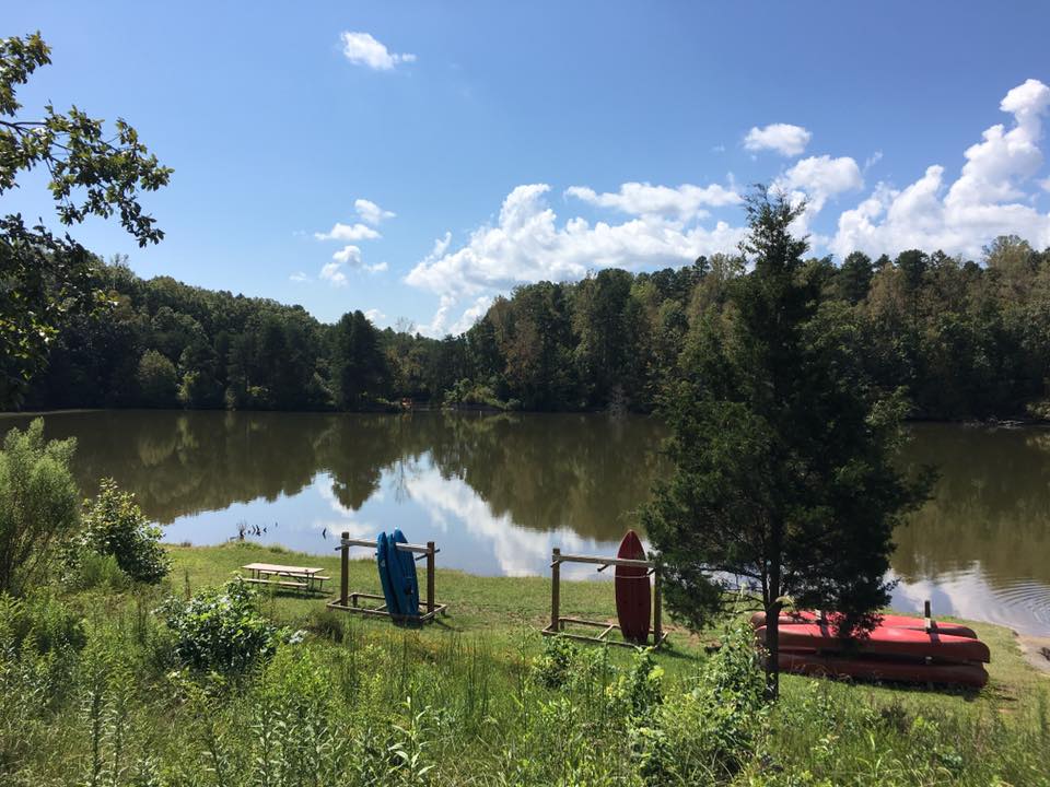 A calm lake surrounded by trees with a few canoes and kayaks on a rack near the shore under a clear blue sky.