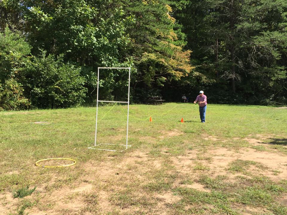 A person holding a string stands on a grassy field next to an empty frame, a hula hoop on the ground, and two orange cones. Trees and a picnic table are in the background.