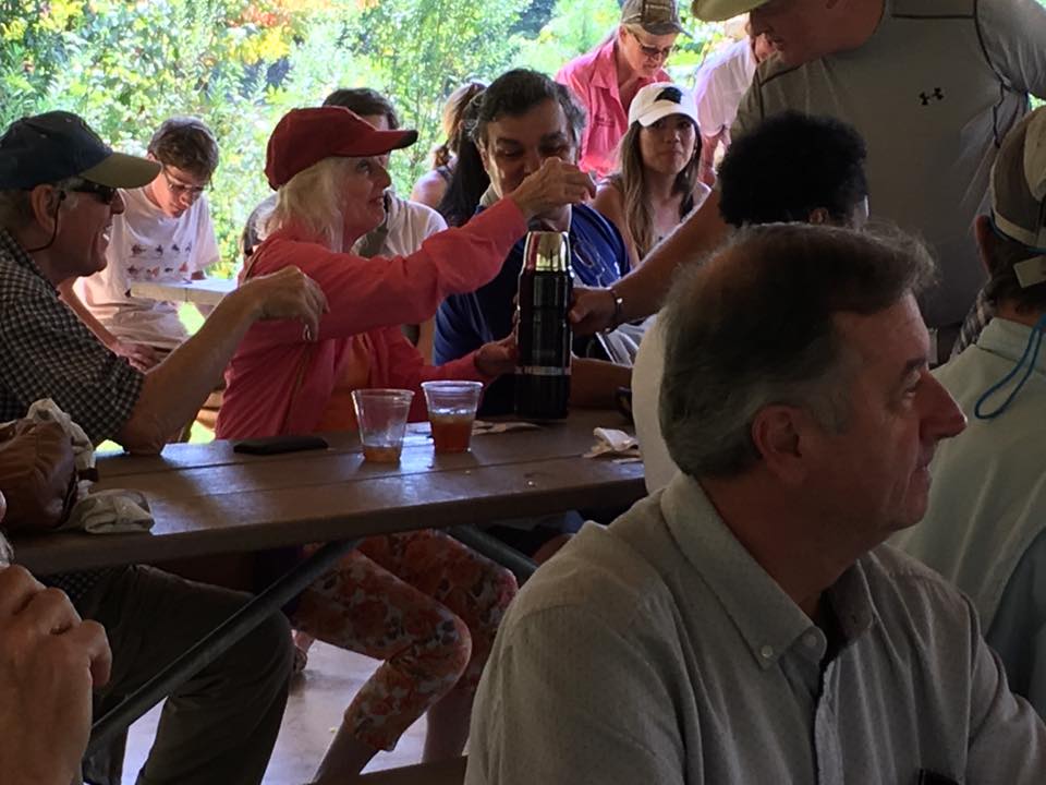 A group of people sits at a picnic bench under a gazebo, sharing drinks and conversing. Trees and foliage are in the background.