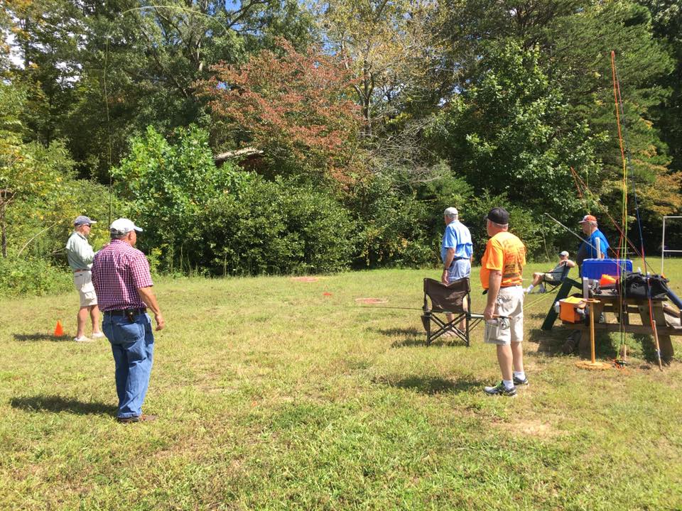 A group of five men stand in a grassy clearing surrounded by trees; one man sits while others appear to be engaged in conversation. Outdoor equipment is visible nearby.
