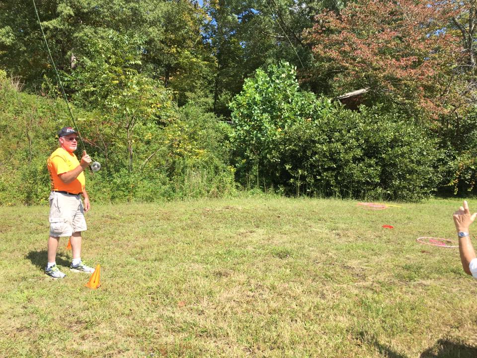 A person in an orange shirt stands on grass, holding a fishing rod, with another person taking a photo of them. Trees and bushes are in the background.