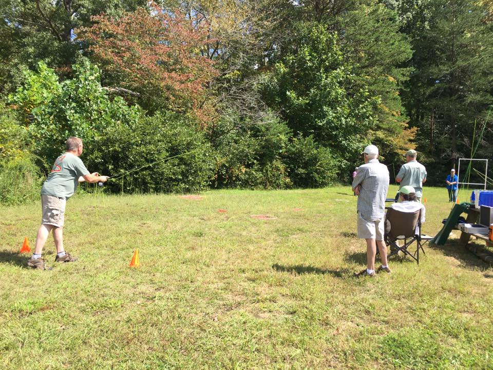 A person practices fly fishing casting on a grassy field with cones marking the area. Several individuals look on, and trees are in the background.