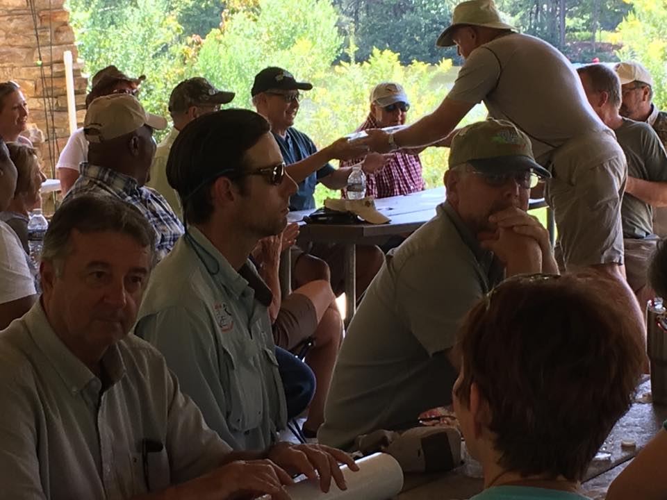 A group of people sit at picnic tables under a pavilion, some wearing hats and sunglasses. A man in a hat is handing something to another seated man. Trees and greenery are visible in the background.