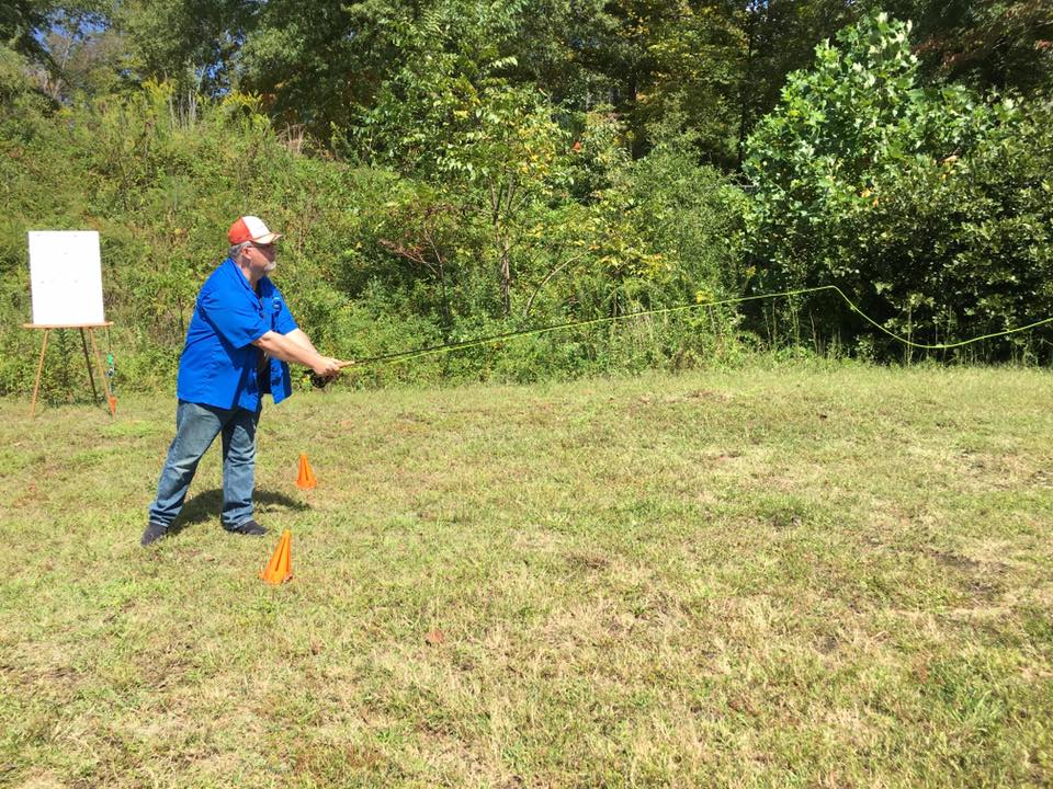A man in a blue shirt and jeans practices fly fishing on a grassy field with trees in the background. Two orange cones are placed near him.