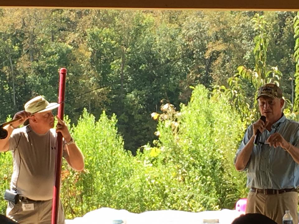 Two men stand outdoors; one holds a long red pipe, while the other speaks into a microphone. They are surrounded by greenery and are under a shelter.