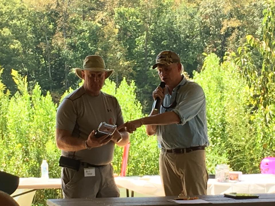 Two men outdoors: one is holding and looking at a device, and the other is speaking into a microphone while pointing at the device. Trees and picnic tables are in the background.