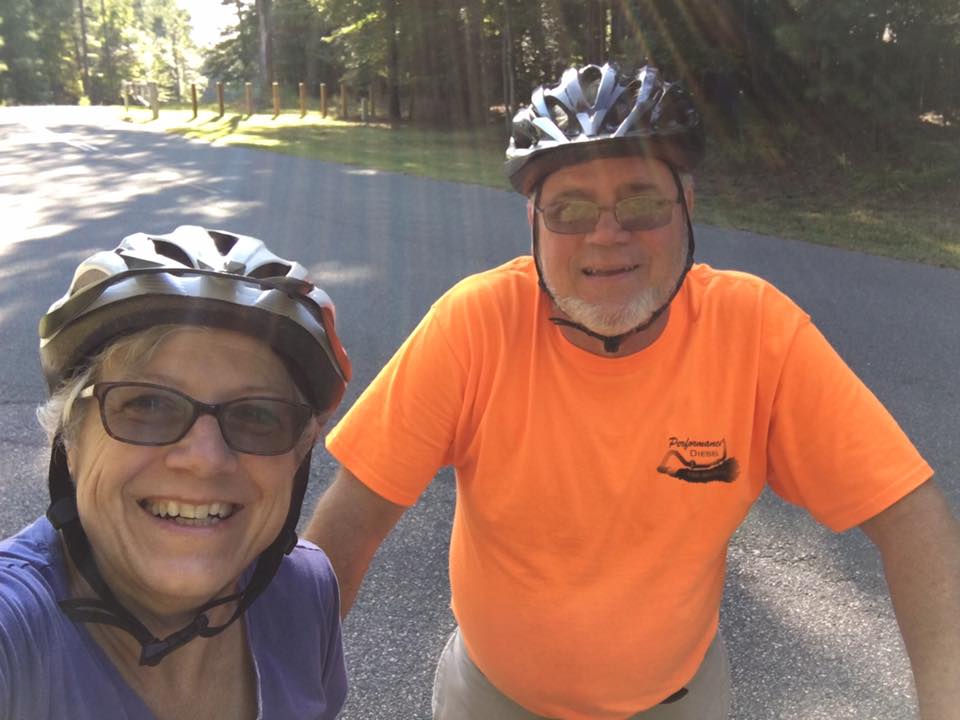 Two people wearing helmets and glasses smile at the camera while standing on a paved road surrounded by trees.