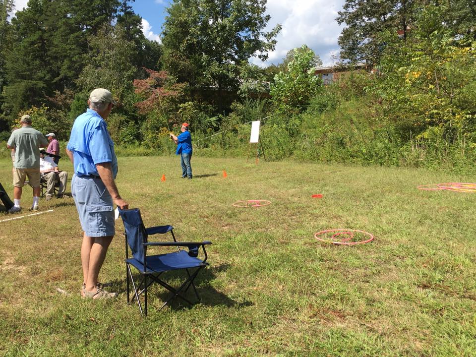 People are playing a lawn game involving rings and poles in a grassy field with trees in the background. One man is standing near a foldable chair.