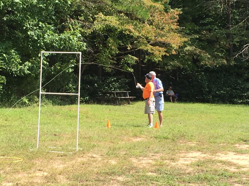 Two people are practicing fly casting in a grassy area with trees in the background. Orange cones and a metal frame are set up on the ground. Another person is seated in the distance.