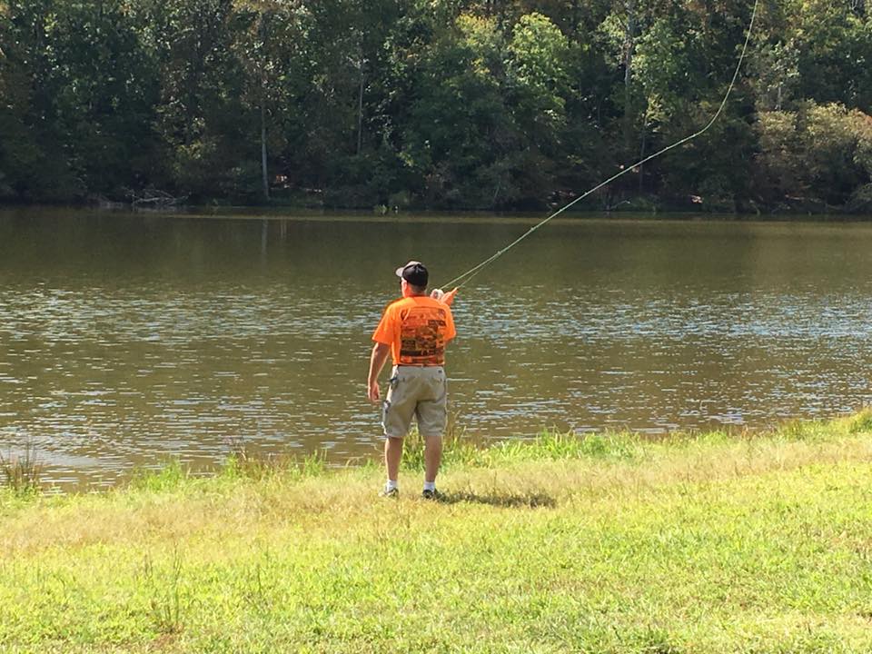 A person in an orange shirt is casting a fishing line into a calm lake, surrounded by grass and trees.