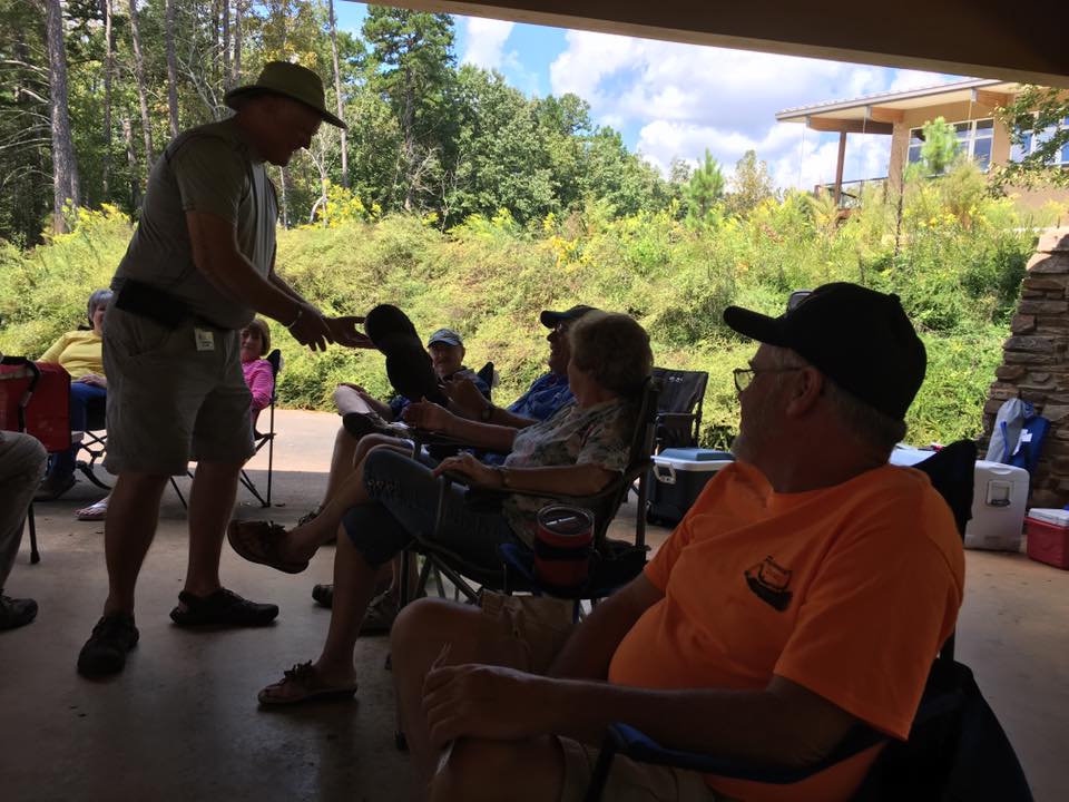 A group of people sit in chairs under a shaded patio, engaged in conversation. A man with a hat appears to hand something to a woman, while others are seated nearby. Greenery and a building are in the background.