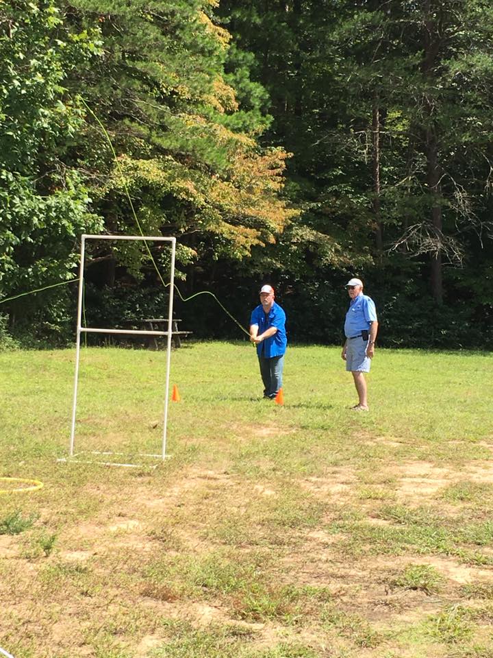 Two people practicing fly casting on a grassy field with trees in the background. One person is casting with a rod, while the other stands nearby watching. A practice target with a frame is visible.