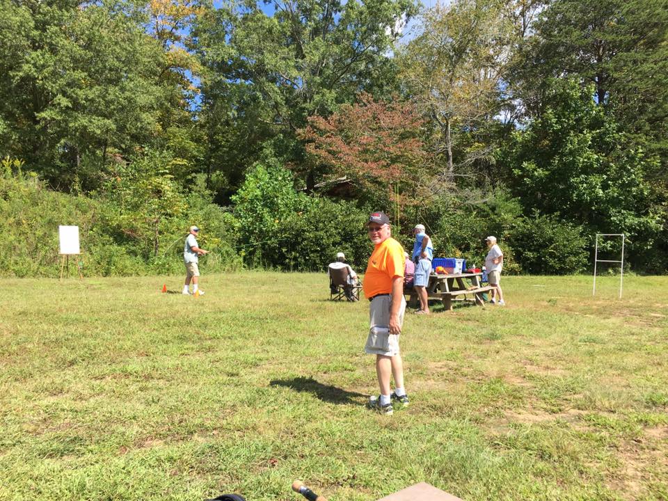 A group of people are outdoors in a grassy area with trees in the background, engaging in an activity with equipment. A man in an orange shirt stands in the foreground.